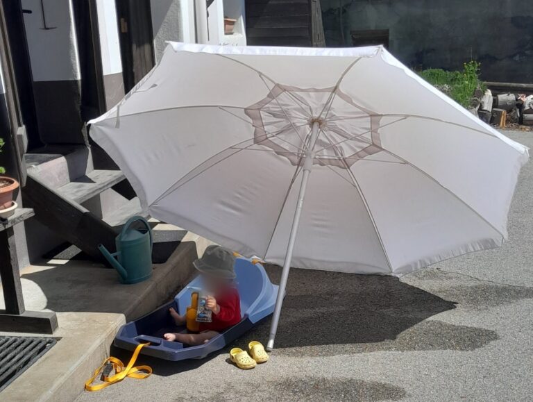 Child sitting in a blue plastic wagon under a large white patio umbrella on a sidewalk, with a yellow sandal nearby and a watering can on the steps nearby.